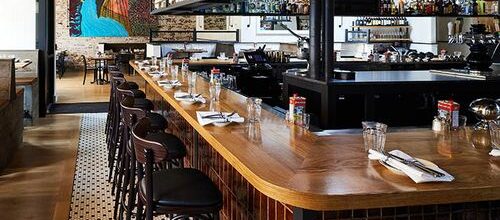 Long curved wooden bar set for dining with barstools, glasses, and colorful artwork on the back wall.