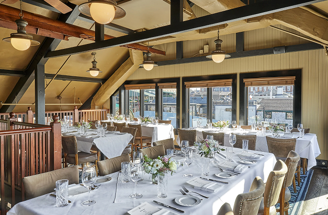 Large dining setup with long tables, white tablecloths, and flowers in a high-ceilinged loft with wooden beams and large windows overlooking a marina.