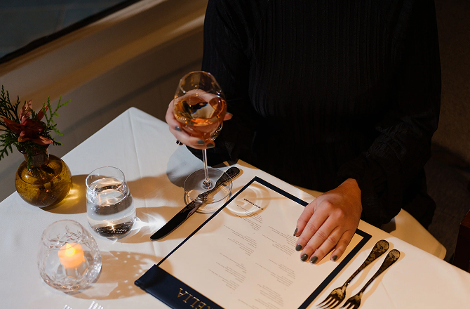 A diner holding a glass of wine while looking over a restaurant menu set on a white table.