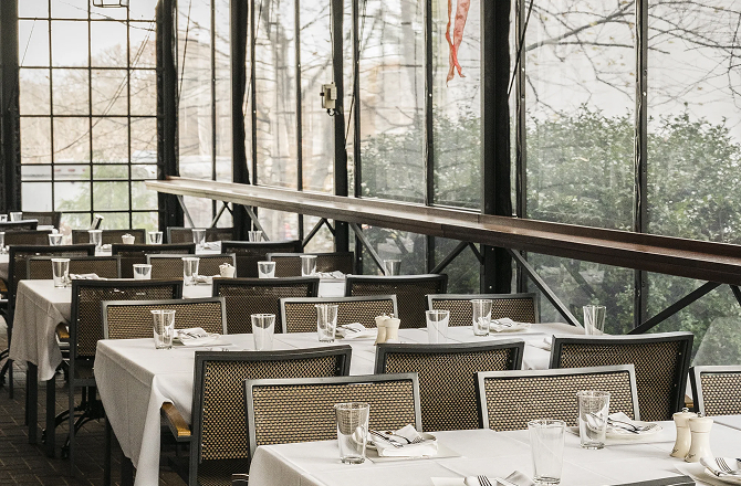 Enclosed patio dining area with long tables set with white tablecloths, glassware, and woven-back chairs