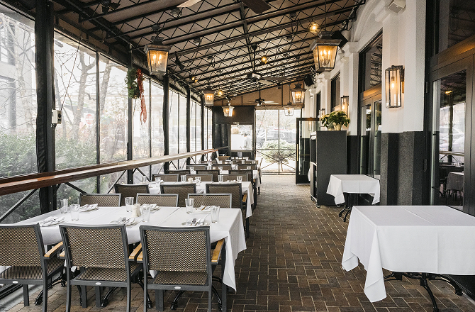 Enclosed patio dining area with long tables set with white tablecloths, glassware, and woven-back chairs