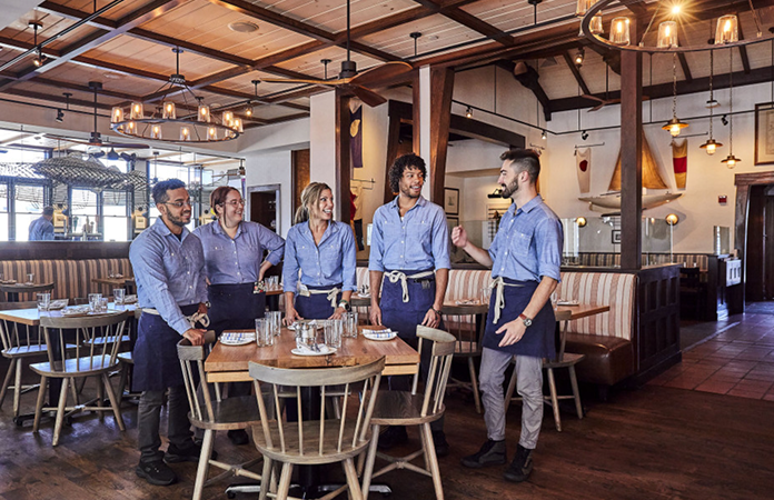 Five restaurant staff members wearing matching blue shirts and aprons standing together in a dining room.