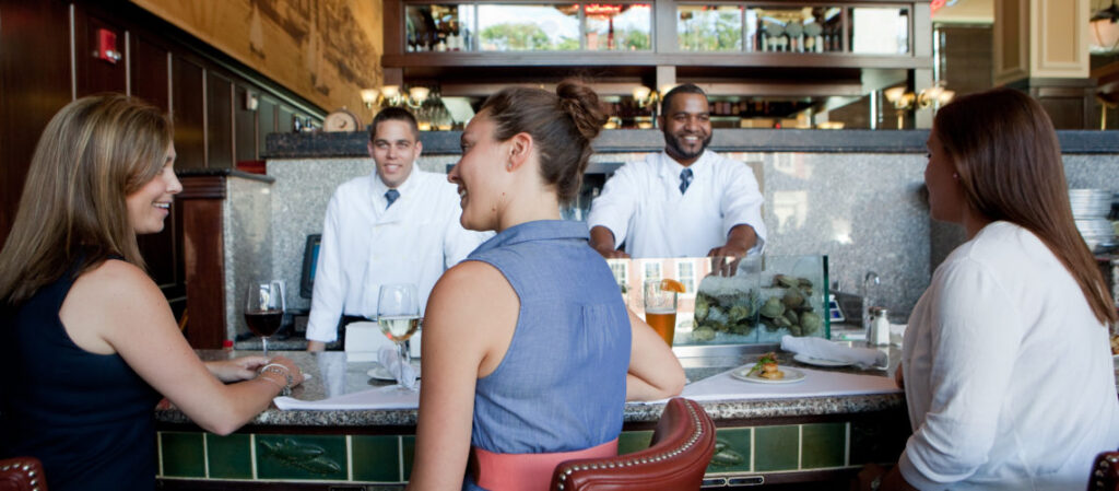 Three women sitting at a bar, chatting with two staff members behind the counter.