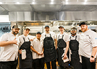 A group of chefs and kitchen staff wearing aprons standing together inside a commercial kitchen.