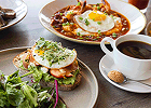 A breakfast spread featuring avocado toast topped with a fried egg, a skillet dish, and a white mug of coffee on a wooden table.