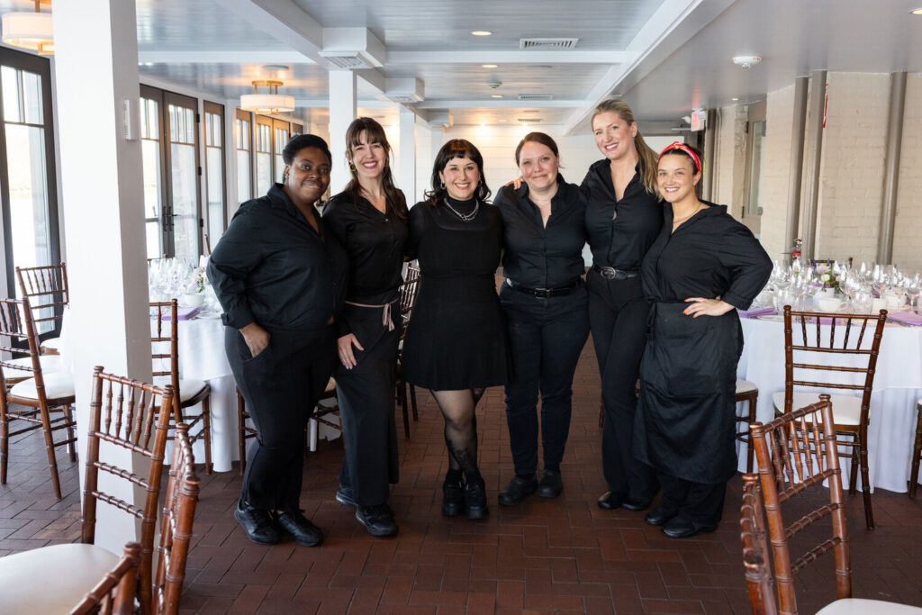 Six women dressed in black attire posing and smiling together inside a dining room set for an event.