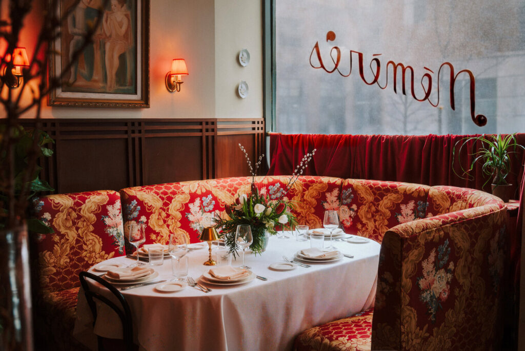 Intimate dining corner featuring a curved red floral patterned booth, a white-linen-covered dining table set with wine glasses and floral arrangements, and red wall sconce lighting.
