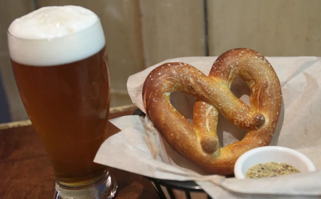 Tall glass of beer next to a large salted pretzel in a basket with a bowl of grain mustard.