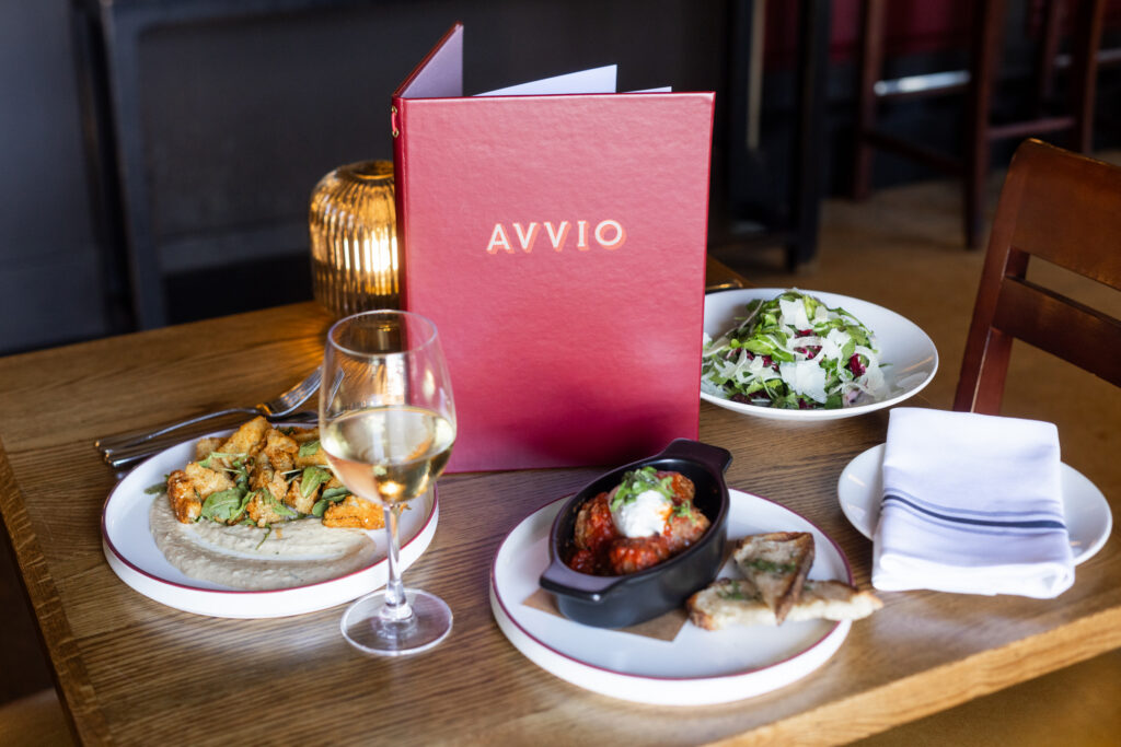 Red Avvio menu, glass of white wine, salad, meatballs, and appetizer on a wooden table.
