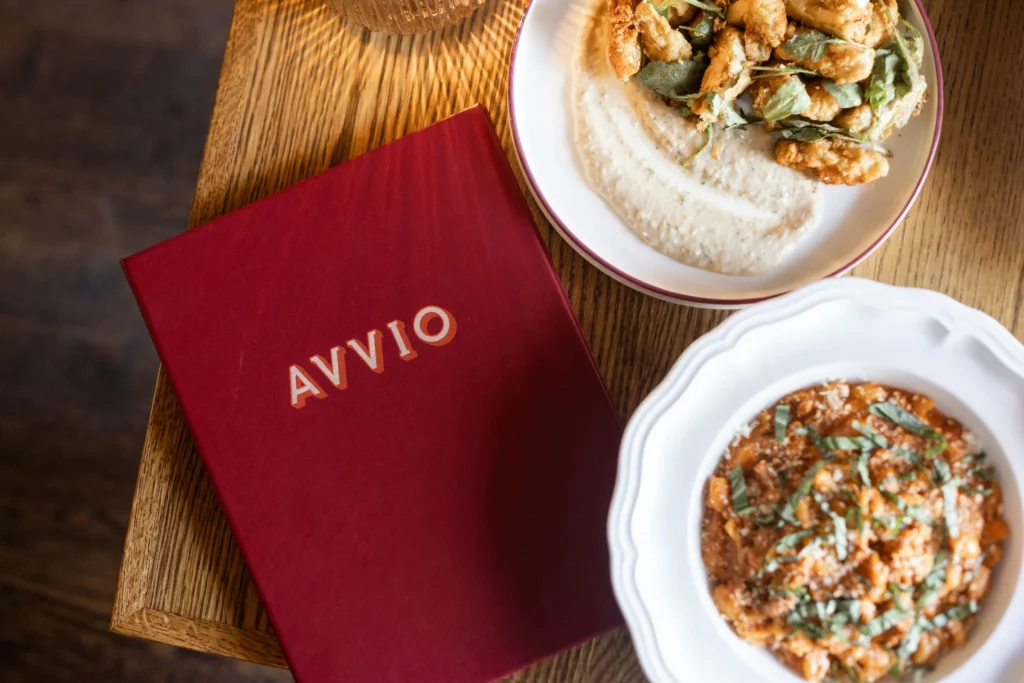 A red Avvio menu sitting on a wooden table alongside plated pasta and appetizer dishes.