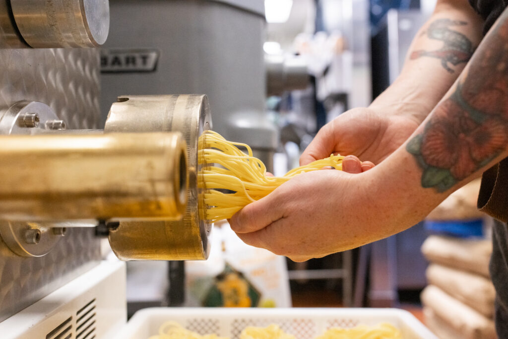 Close up of hands catching fresh spaghetti noodles coming out of a metal pasta extruder.
