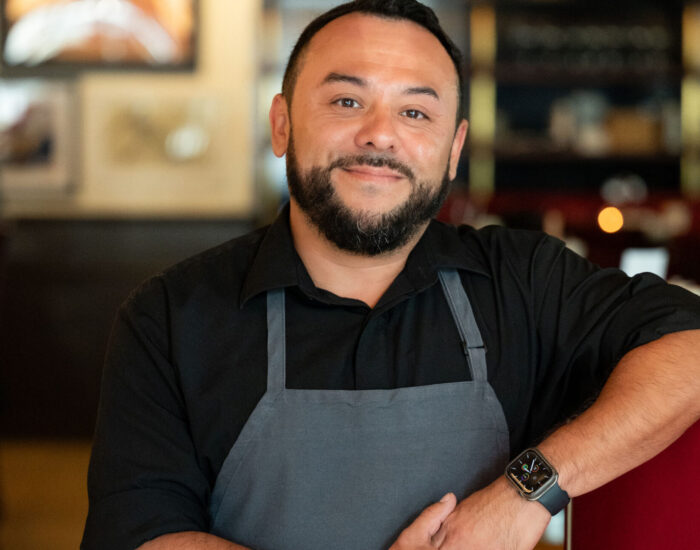 Smiling man with dark hair and a beard wearing a black collared shirt and dark grey apron.