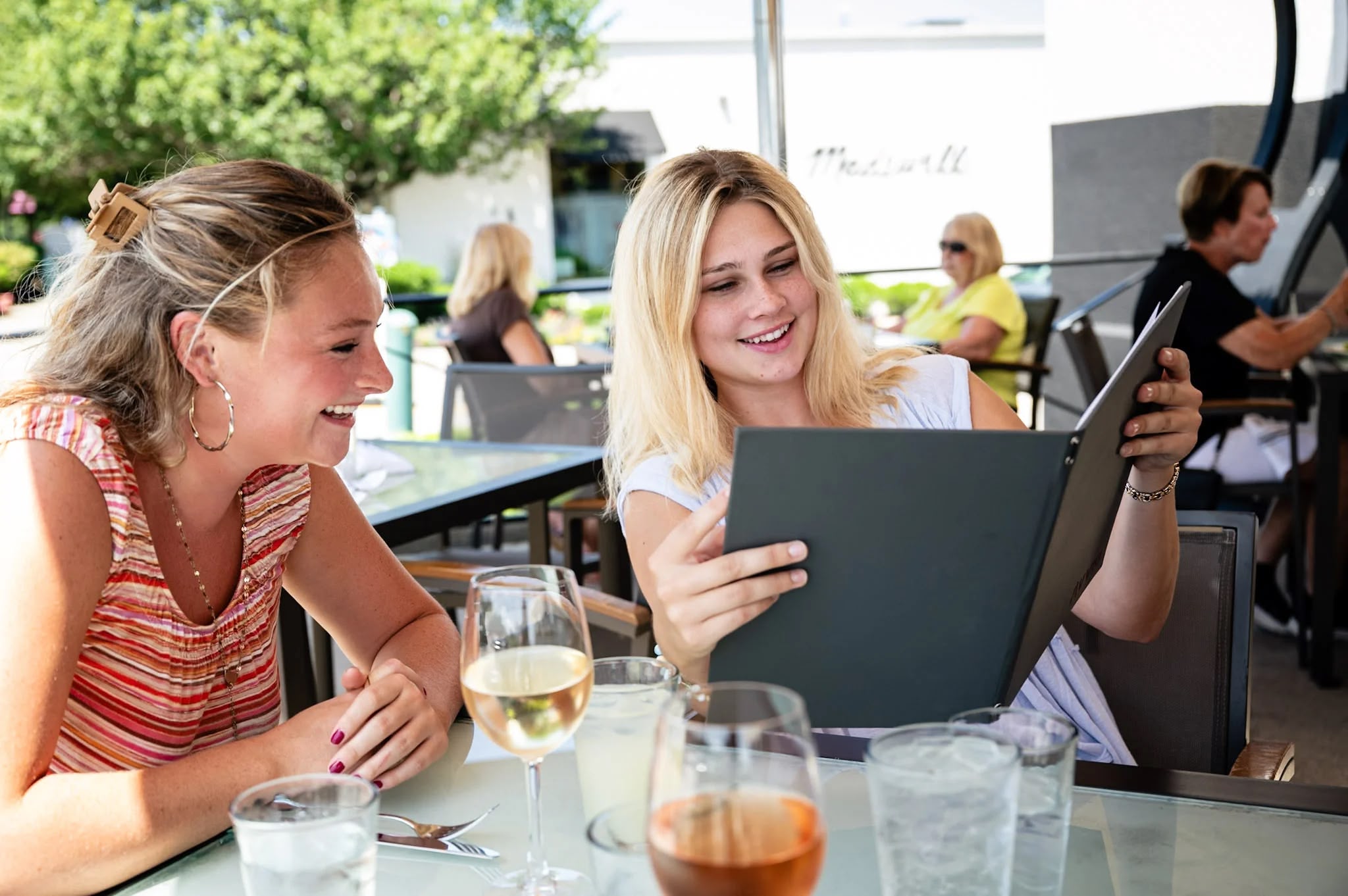 Two young women smiling while looking at a menu at an outdoor restaurant table with drinks.