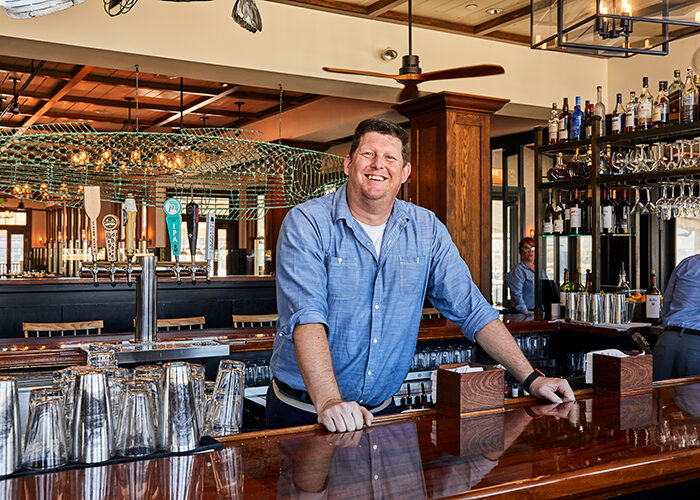 Smiling man in a denim shirt resting his hands on a polished wooden bar inside a restaurant.
