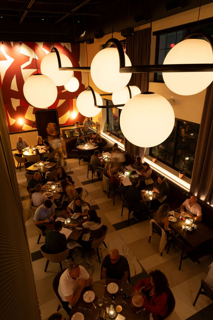 High angle view of a bustling restaurant filled with seated diners under large globe pendant lights and a red patterned wall art.