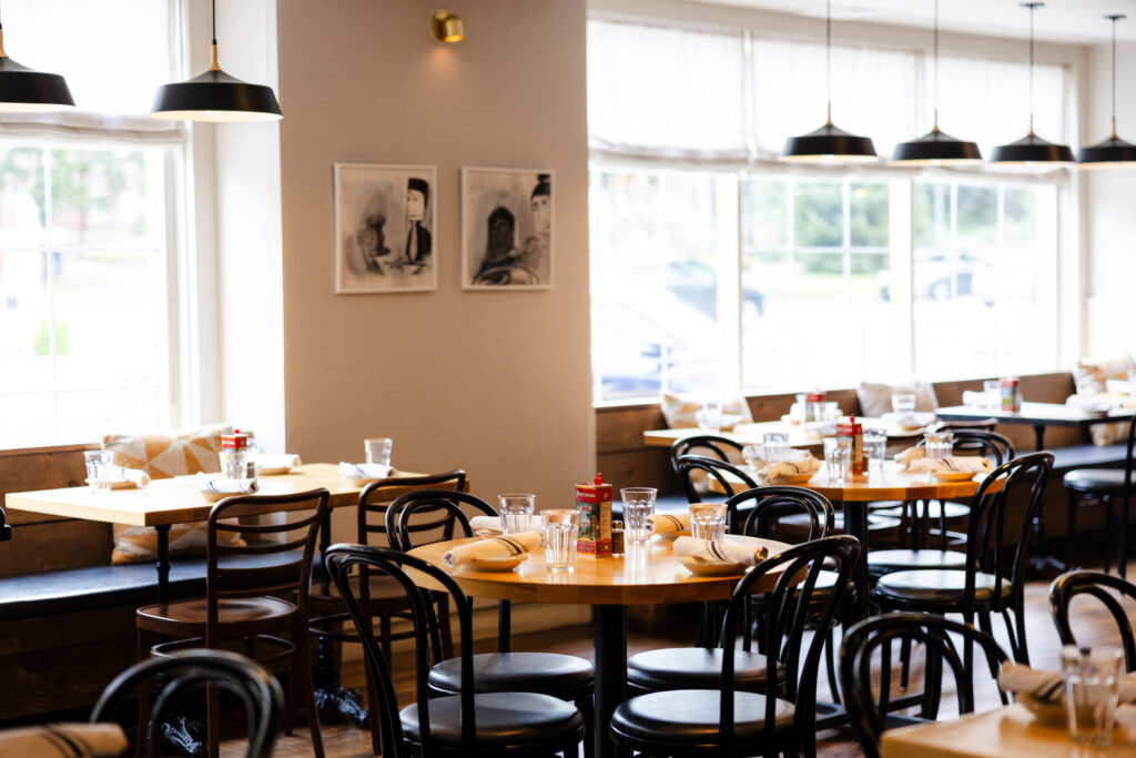 Bright dining area at with light wooden tables, black chairs, artwork on beige walls, and black pendant lights.