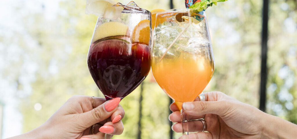 Two hands toasting with colourful cocktails in wine glasses garnished with fruit slices outdoors.