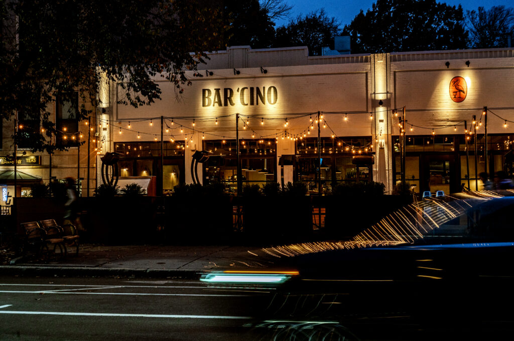 Exterior of Bar Chino at night lit by string lights with light trails from passing car traffic in the foreground.