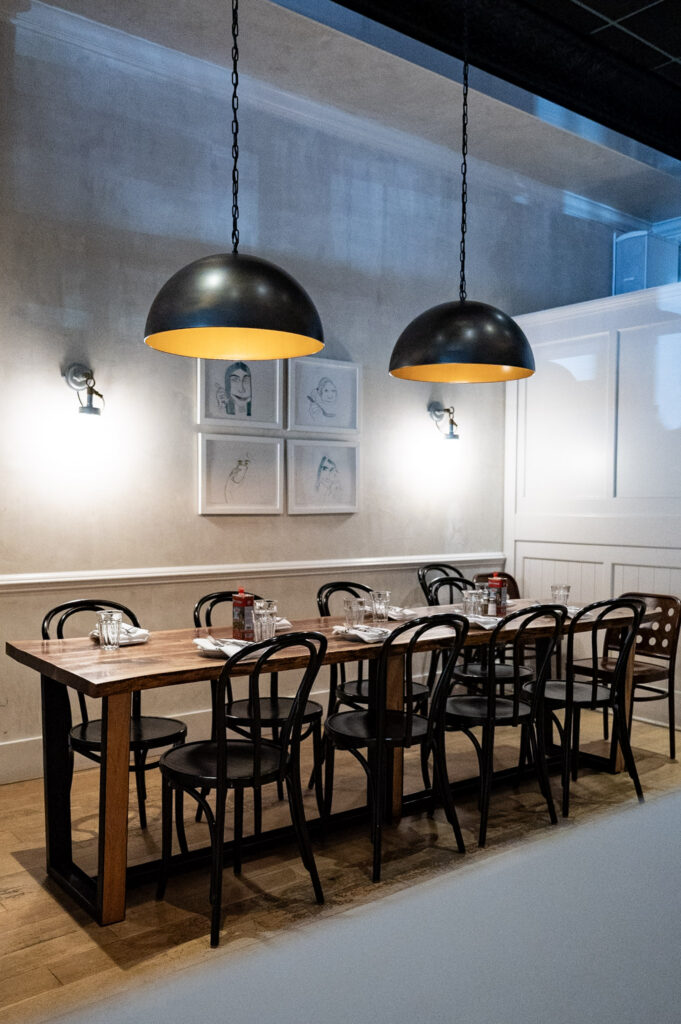 Long wooden dining table set with black chairs, place settings, and two black dome pendant lights.