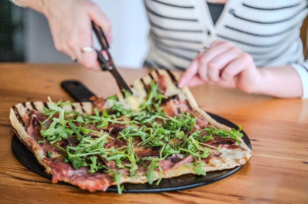 Person using shears to cut a flatbread pizza topped with prosciutto and fresh arugula on a black tray