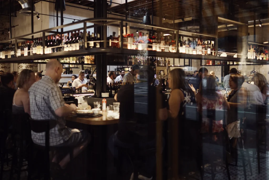 Patrons sitting at a busy restaurant bar seen through a window.