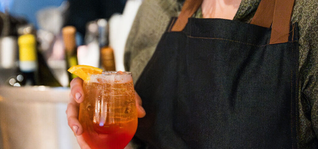 A person in a dark apron holding an orange cocktail garnished with an orange slice.