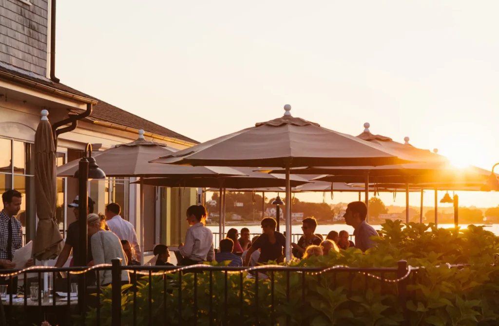 Guests dining on a patio under umbrellas overlooking the water at sunset