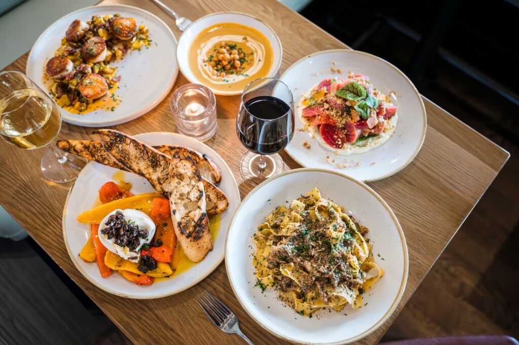 Overhead view of a wooden table spread with multiple plates of gourmet food pasta soup seafood and glasses of wine.