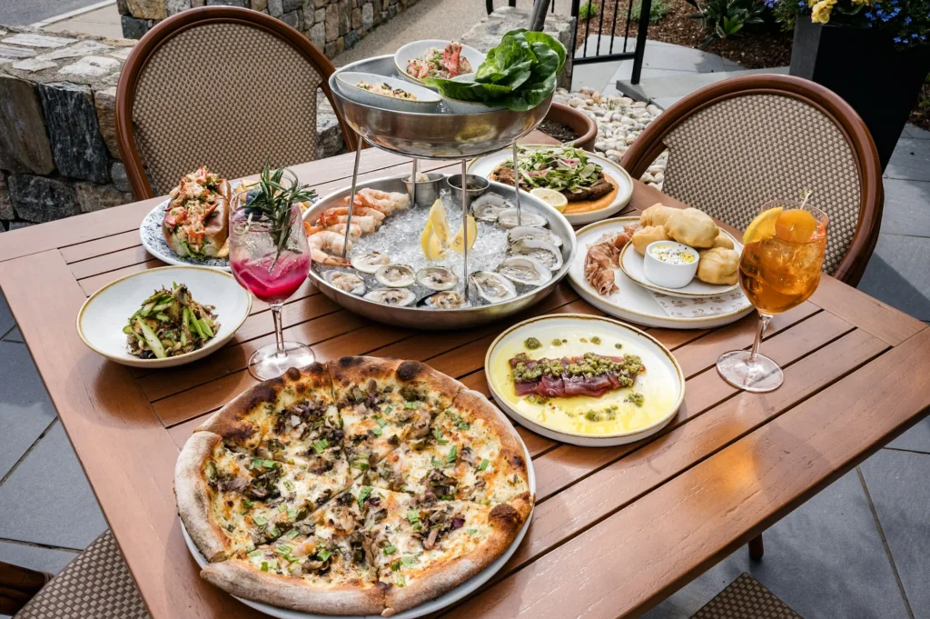 A wooden table filled with various seafood dishes, a pizza, and colorful cocktails outdoors.