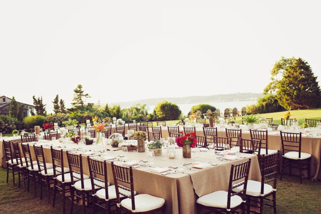 Outdoor wedding reception tables set up on a lawn near the water with white chairs and floral centerpieces.