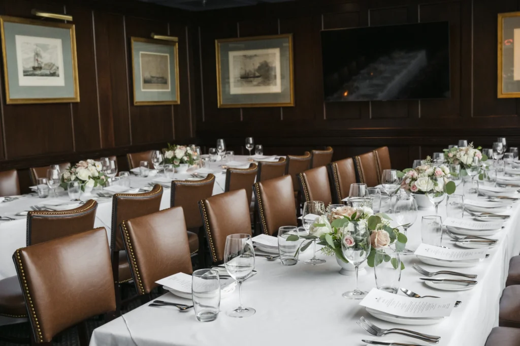Long banquet tables set with white tablecloths, brown leather chairs, floral centerpieces, glassware, and framed nautical art on dark wood walls.