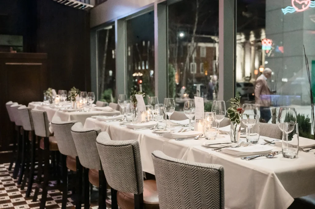 Long dining table set for an event with white linens, wine glasses, and candles beside large windows.