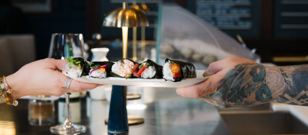 Two hands passing a white plate filled with sushi hand rolls.