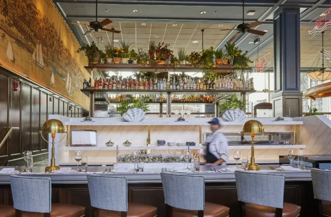 A chef works behind a marble raw bar counter lined with seating, framed by brass lamps and potted plants under a high ceiling.