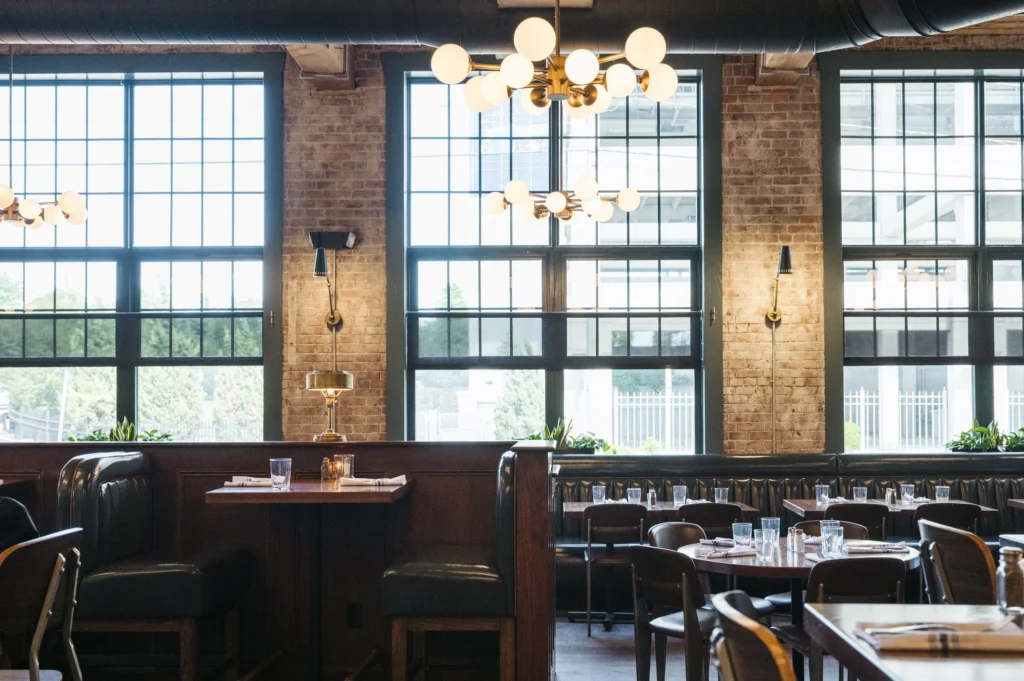 Restaurant dining room with large grid windows, brick walls, dark wood booths, tables, and a modern globe chandelier.
