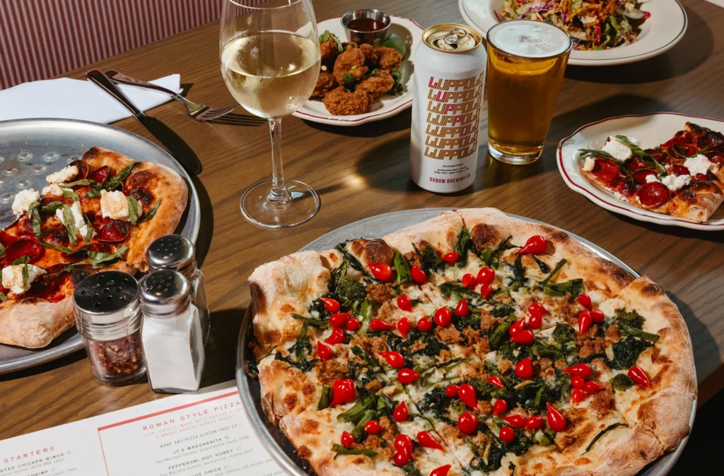 A dining table filled with pizzas, sides, beer, and a glass of white wine set beside a restaurant menu.
