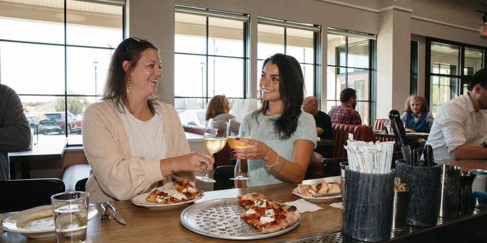 Two women smiling and clinking white wine glasses at a restaurant table with pizzas in front of them.
