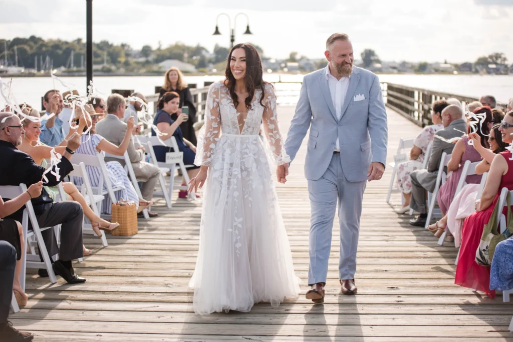 Bride and groom walk down a wooden dock aisle holding hands, surrounded by seated wedding guests outdoors.