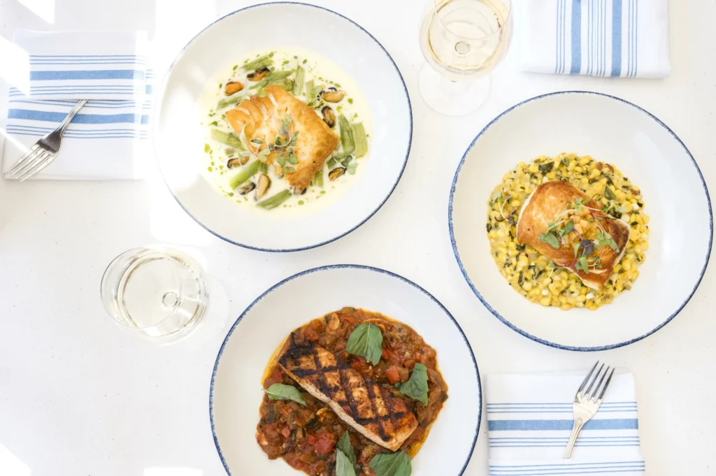 An overhead view of three plated seafood entrees served on blue rimmed plates alongside striped napkins and silverware.
