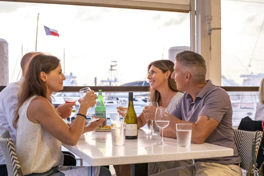 Four friends enjoying wine and conversation at a sunny waterfront restaurant with boats visible through the window.
