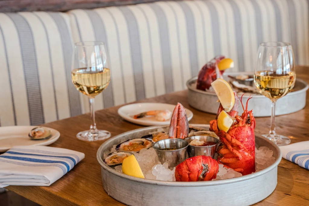 A seafood platter featuring a whole lobster and oysters on ice served with two glasses of white wine on a wooden table.