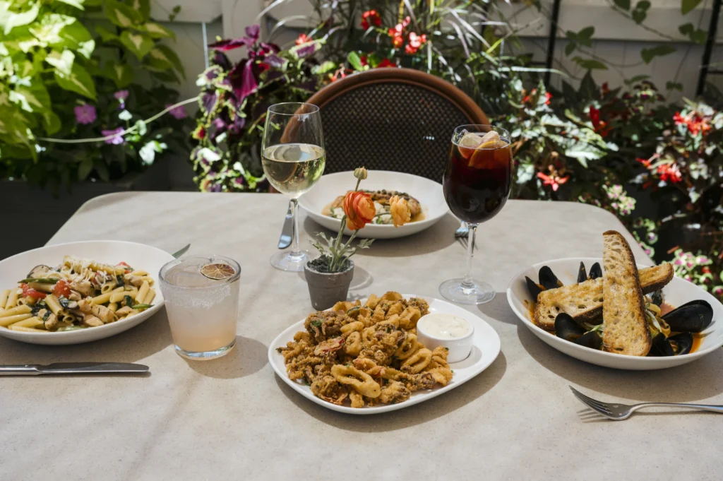 An outdoor restaurant table set with various Italian dishes including calamari, pasta, mussels, white wine, and sangria.