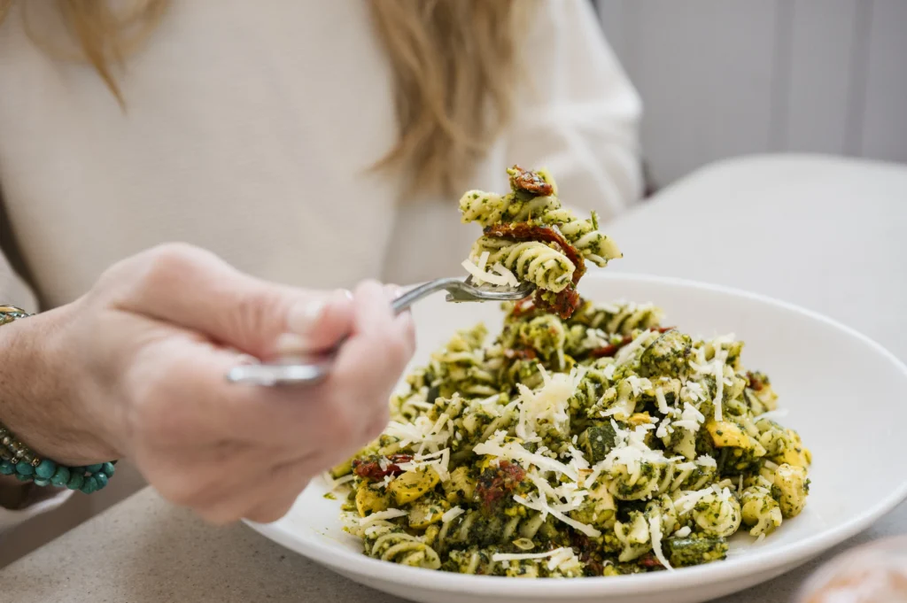 A person using a fork to lift a bite of pesto fusilli pasta topped with grated cheese from a plate.
