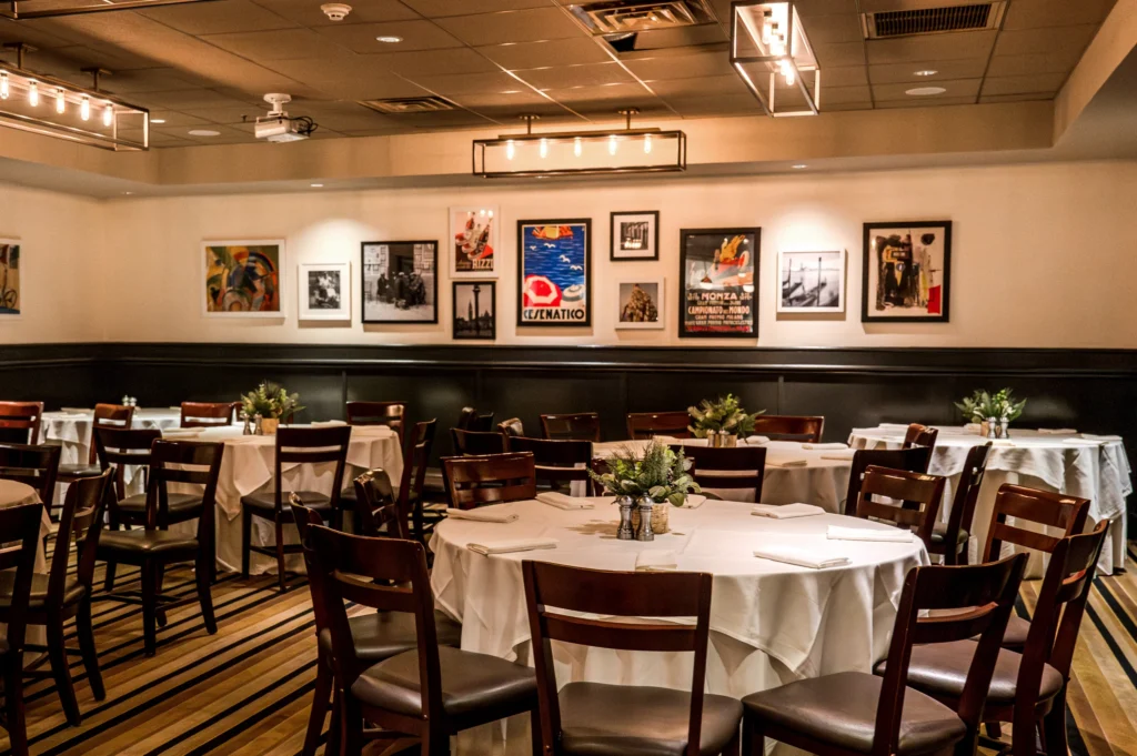 A dining room with round tables, white tablecloths, dark wooden chairs, and framed artwork on light walls under warm lighting.