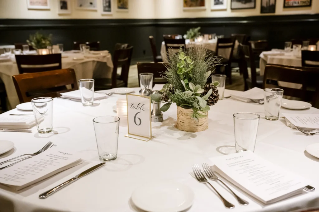 A dining table set with white tablecloths, glassware, silverware, menus, a table number sign, and a centerpiece with pinecones and greenery.