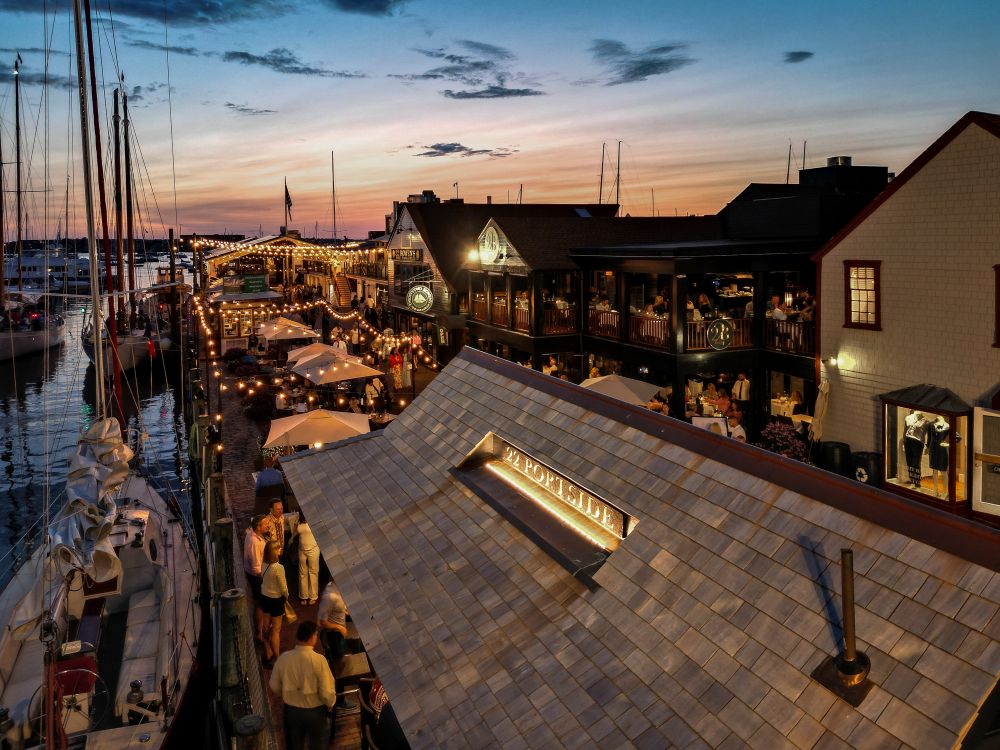 An aerial evening view of a bustling coastal harbor lined with sailboats, illuminated restaurants, and people walking along a wooden boardwalk.