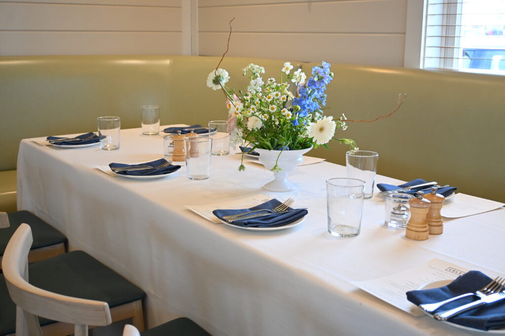 Long table with a white tablecloth, navy napkins, glassware, and a centerpiece of white and blue flowers next to a booth seating area.