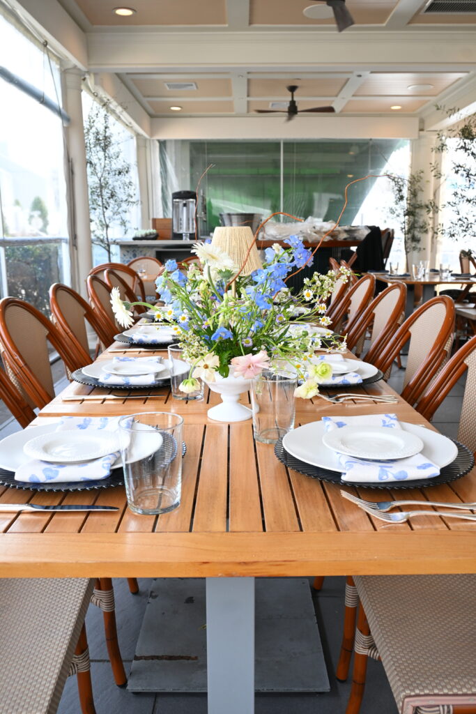 Long wooden patio dining table set with white and floral plates, blue floral napkins, woven chairs, and a floral centerpiece.