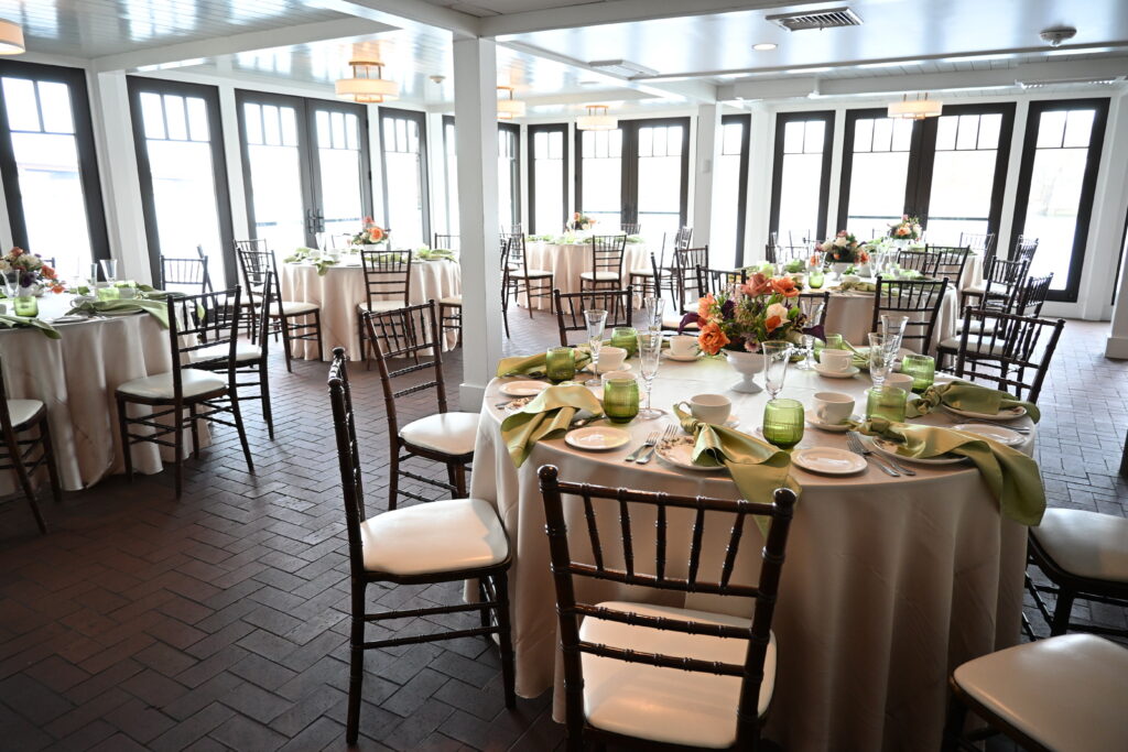Spacious indoor event room set up with round dining tables, beige tablecloths, green napkins, chiavari chairs, and large glass windows.