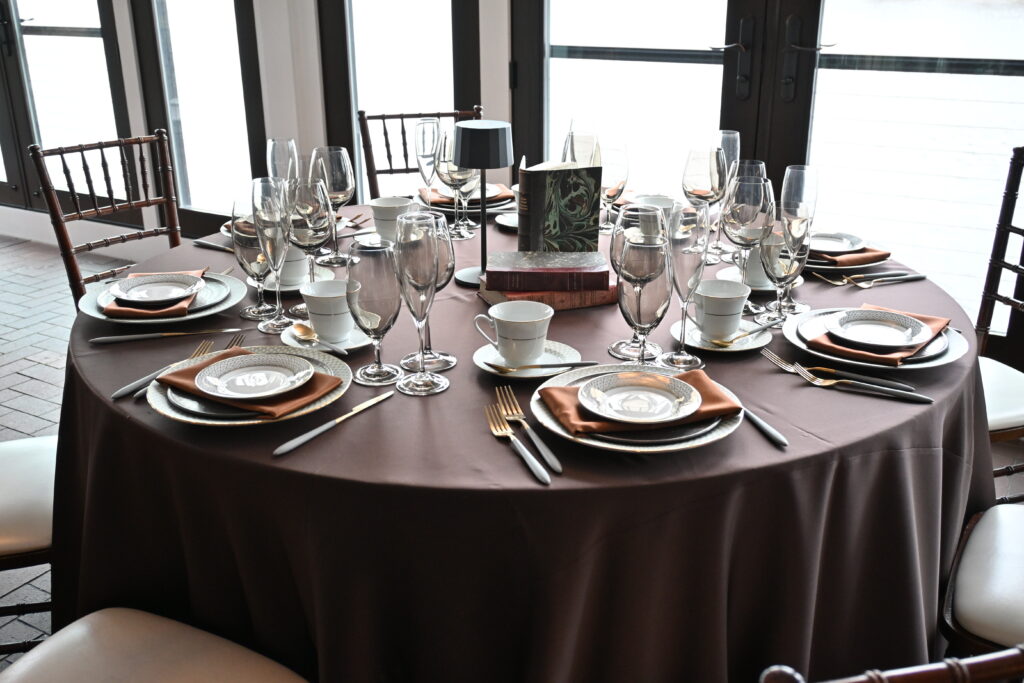 A round dining table set with brown tablecloth, gold silverware, fine china, wine glasses, and book centerpiece.